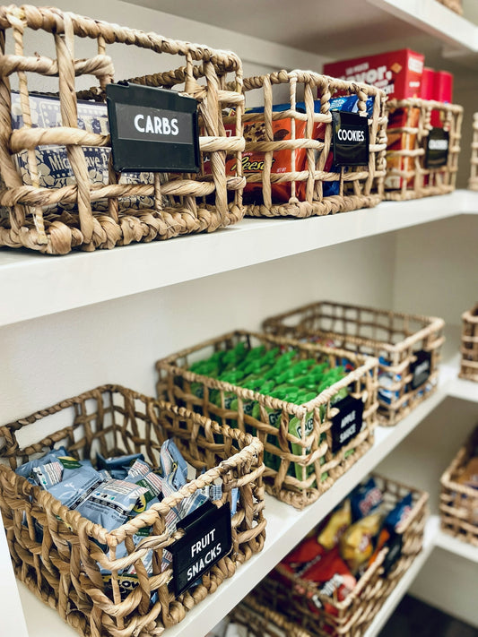 Woven baskets with labels on white pantry shelves filled with various snacks